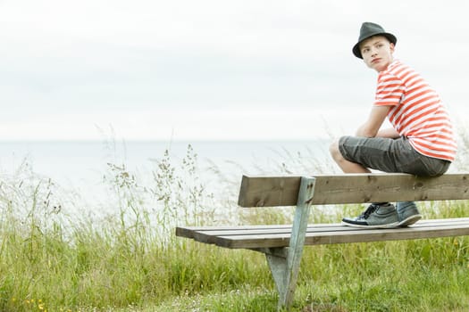 Rear view of teenager in striped shirt and hat sitting on top of wooden bench in front of ocean looking over his shoulder