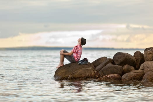 Young man enjoying an ocean sunset sitting barefoot on rocks at the edge of the sea wearing a trendy hat looking up into the air, side view with copy space