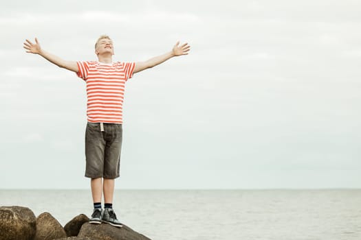 Single young short haired teenager in striped shirt with big smile and wide open arms standing on large rocks near water with copy space