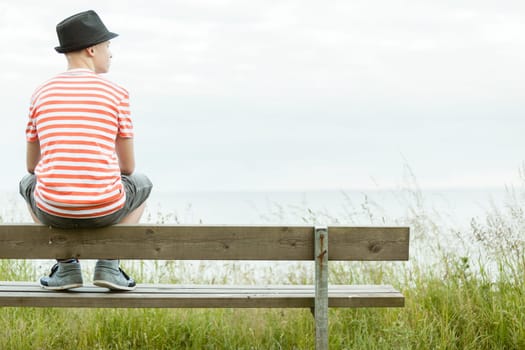 Teenage boy in a trendy hat sitting on a rustic wooden bench overlooking the sea with his back to the camera and copy space