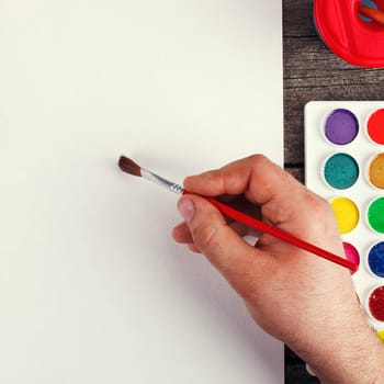 hand with brush painting with watercolor  and blank white paper sheet  on vintage wooden background