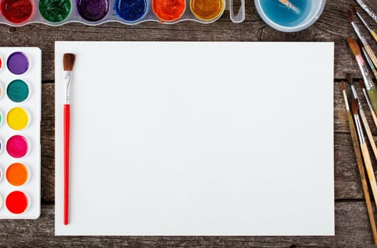 Set of watercolor paints,  brushes for painting and blank white paper sheet of sketchbook on vintage wooden background. Top view.