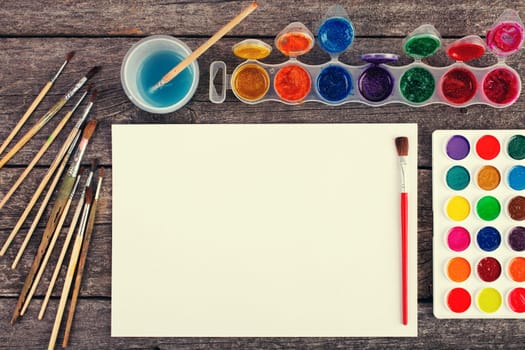 Set of watercolor paints,  brushes for painting and blank white paper sheet of sketchbook on vintage wooden background. Top view.