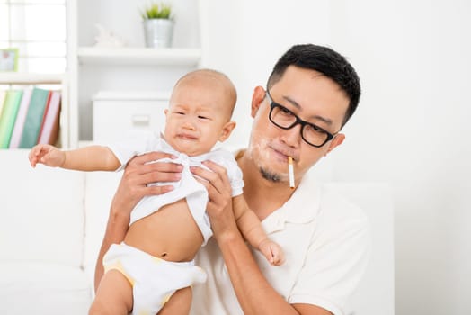 Asian family at home. Bad father smoking while holding baby together. Cigarette with lit and smoke. Unhealthy lifestyle or stop smoking concept photo.