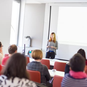 Speaker giving presentation in lecture hall at university. Participants listening to lecture and making notes.