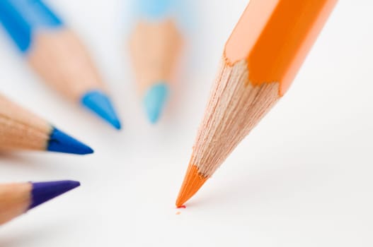 flat-angled detail view of an orange wood colored pencil slanted on paper in front of four flat colored pencils in blue tones,the tips circular to the pen in the foreground pointing on drawing paper