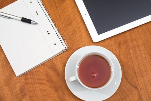 tablet PC, notepad with pen and a cup of tea on wooden table from above