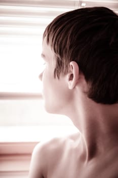 Close up head shot of a shirtless thoughtful young boy staring out of a bright window viewed in profile