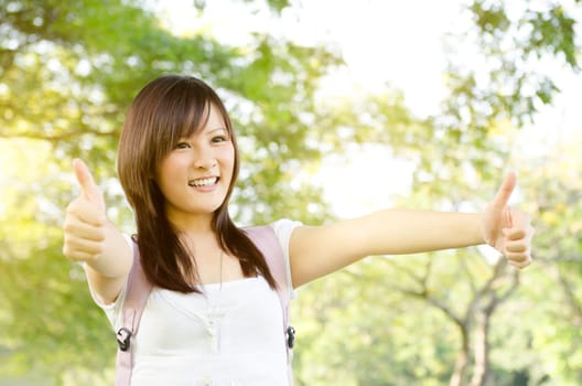 Young Asian university girl student standing on campus lawn, showing thumbs up and smiling.
