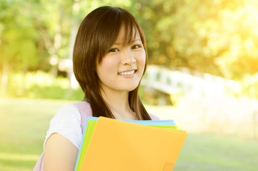 Young Asian university student standing on campus lawn, holding file folder and smiling.