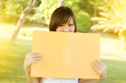 Young Asian college girl student standing on campus lawn, smiling and covered face by file folder.