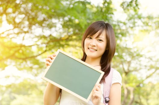 Young Asian university girl student standing on campus lawn, holding a blank chalkboard and smiling.