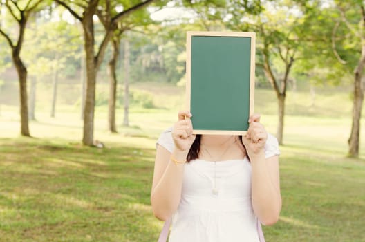 Young Asian college girl student standing on campus lawn, hands holding a blank chalkboard and covered face.