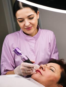 Permanent makeup artist tattoos a beautiful woman over 50 years old in a beauty salon on the client's face. Vertical