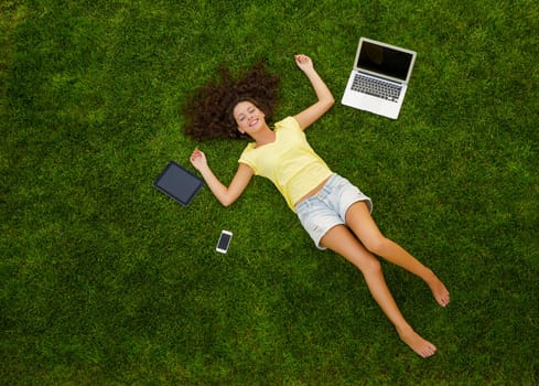 Beautiful and happy young woman lying on the grass surrounded by technology devices