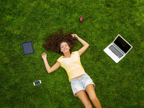 Beautiful and happy young woman lying on the grass surrounded by technology devices