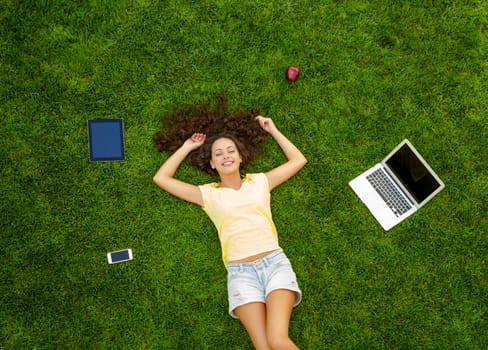 Beautiful and happy young woman lying on the grass surrounded by technology devices