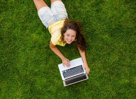 Beautiful young woman lying on the grass and working with a laptop