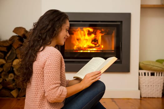 Beautiful woman reading a book at the warmth of the fireplace