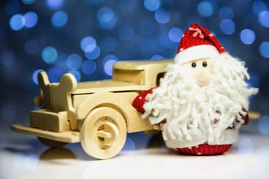 Santa Claus or Father Frost with old vintage wooden automobile on christmas lights background. Main focus of image on Santa Claus and selective on turned left wood car