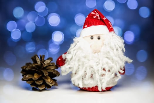 Santa Claus or Father Frost and pine cone with Christmas lights at background on white surface. Main focus of image on Santa Claus and selective on pine cone