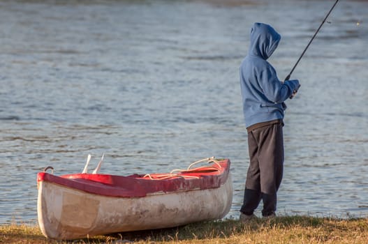 Boy, standing next to canoe on the banks of a river, with fishing rod in hand, busy fishing.