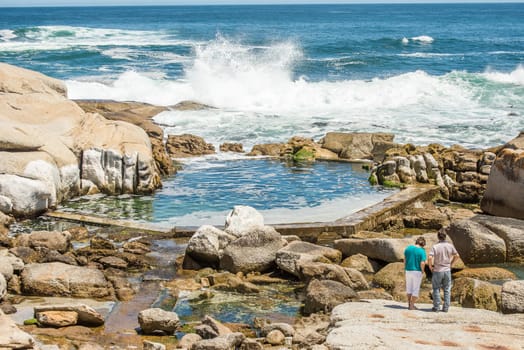 Two people standing on the rocks at Sea Point, Cape Town, near a man made tidal pool, watching the waves crashing in to the rocks of the shore.