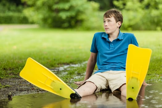 Daydreaming male teenager wearing shorts and diving fins sitting back in large backyard puddle