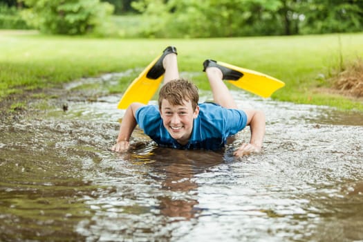 Happy teenager in diving fins laughs as he tries to swim in large shallow muddy backyard puddle