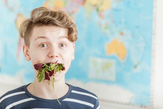 Humorous Portrait of Boy with Wavy Blond Hair Looking Surprised with Mouthful of Mixed Lettuce Salad Greens in Classroom with World Map in Background with Copy Space