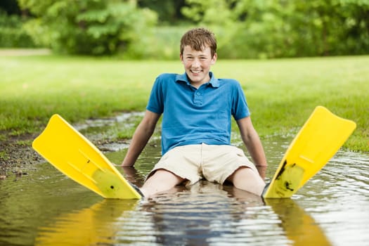 Laughing male teenager in blue shirt, shorts and diving fins laying back in large backyard puddle