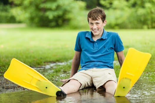 Happy soaked male teenager wearing shorts and diving fins sitting back in large backyard puddle