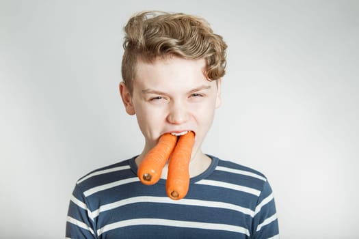 Head and Shoulders Portrait of Boy with Wavy Hair with Two Large Carrots Stuffed into Mouth in front of White Studio Background with Copy Space - Health and Nutrition Humorous Image
