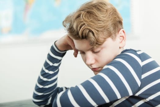 Head and Shoulders Close Up of Boy with Wavy Blond Hair Wearing Striped Shirt Sitting with Head Resting on Hand at Desk in Classroom with World Map in Background