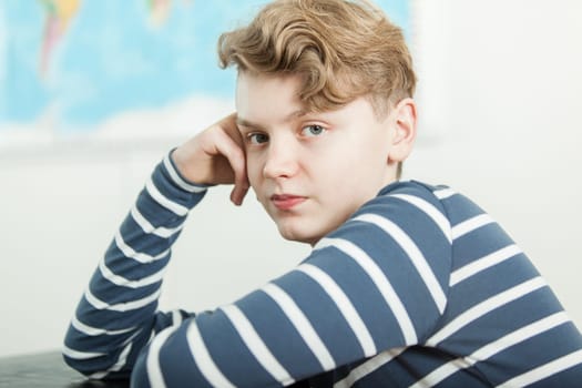 Head and Shoulders Portrait of Boy with Blond Wavy Hair Sitting at Desk with Head Resting on Hand in Classroom with World Map in Background