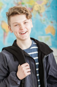 Waist Up Portrait of Smiling Boy with Wavy Blond Hair Standing in Classroom with World Map in Background, Wearing Jacket and Holding Knapsack