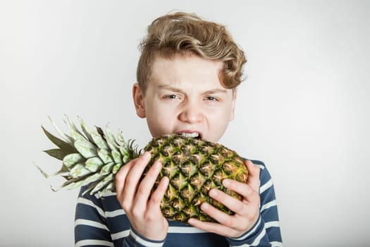 Head and Shoulders of Boy with Wavy Blond Hair Biting into Tough Skin of Spikey Pineapple in Studio with White Background with Copy Space