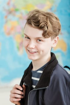 Head and Shoulders Portrait of Smiling Boy with Wavy Blond Hair Standing in Classroom with World Map in Background, Wearing Jacket and Holding Knapsack