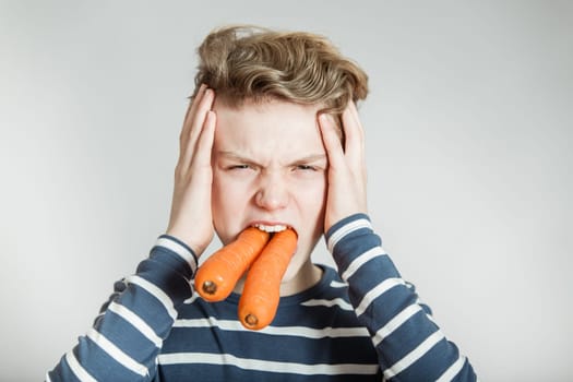 Head and Shoulders Portrait of Boy with Wavy Hair with Two Large Carrots Stuffed in Mouth Holding Head in Pain in front of White Studio Background with Copy Space - Health and Nutrition Humorous Image