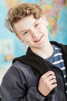 Head and Shoulders Portrait of Smiling Boy with Wavy Blond Hair Standing in Classroom with World Map in Background, Wearing Jacket and Holding Knapsack
