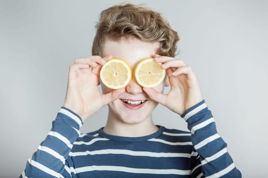 Humorous Nutrition Themed Concept Image of Smiling Boy with Wavy Blond Hair Holding Slices of Lemons Over Eyes in Studio with Gray Background and Copy Space