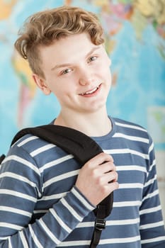 Head and Shoulders Portrait of Smiling Boy with Wavy Blond Hair Standing in Classroom with World Map in Background, Wearing Blue and White Striped Shirt and Holding Knapsack