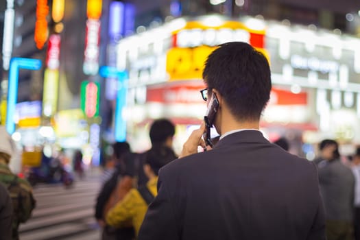 Japanese corporative businessman in suit, after work, talking on cell phone while waiting on crossroad in Kabukicho, entertainment and red-light district in Shinjuku, Tokyo, Japan.