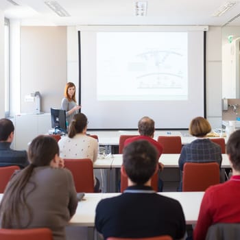 Speaker giving presentation in lecture hall at university. Participants listening to lecture and making notes.