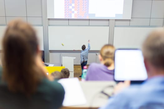 Speaker giving presentation in lecture hall at university. Participants listening to lecture and making notes.