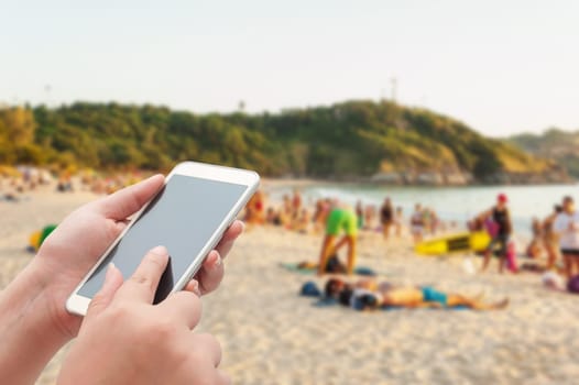 Women using smartphone top on blurred sea and sand beach with some people in evening light - Can be used for display or montage your products