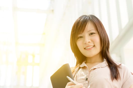 Young Asian businesswoman smiling and holding file folder, standing in an office environment, beautiful golden sunlight at background.
