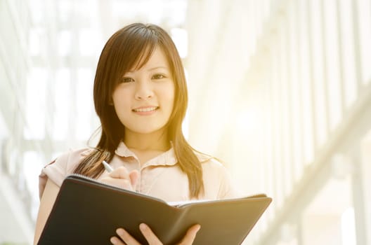 Young Asian business woman smiling and holding file folder, standing in an office environment, natural golden sun light at background.