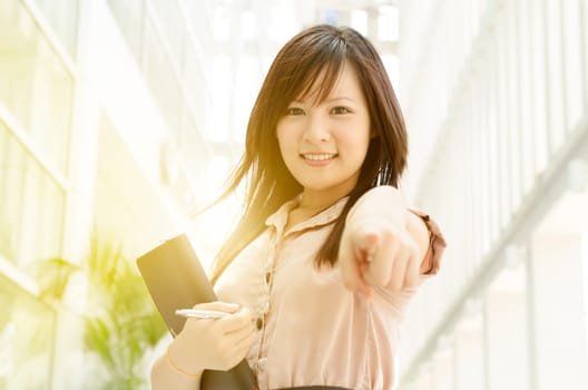 Young Asian business woman smiling and pointing at you, standing in an office environment, natural golden sun light at background.