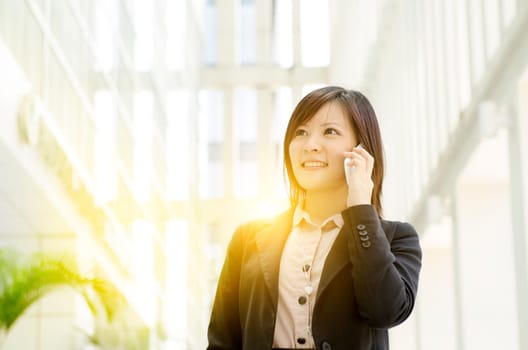 Portrait of a young Asian business woman smiling and talking on smart phone, walking at an office environment, beautiful golden sunlight at background.